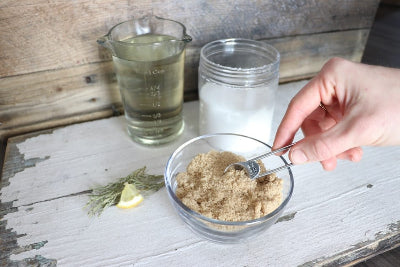 Hand holding a spoon over a bowl of brown powder on a wooden surface with a measuring cup and container in the background.