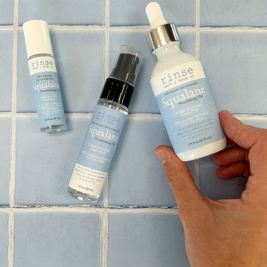Three Rinse Squalane skincare bottles from Body & Soul are arranged on a light blue tiled surface, with a hand holding one of the white dropper bottles.
