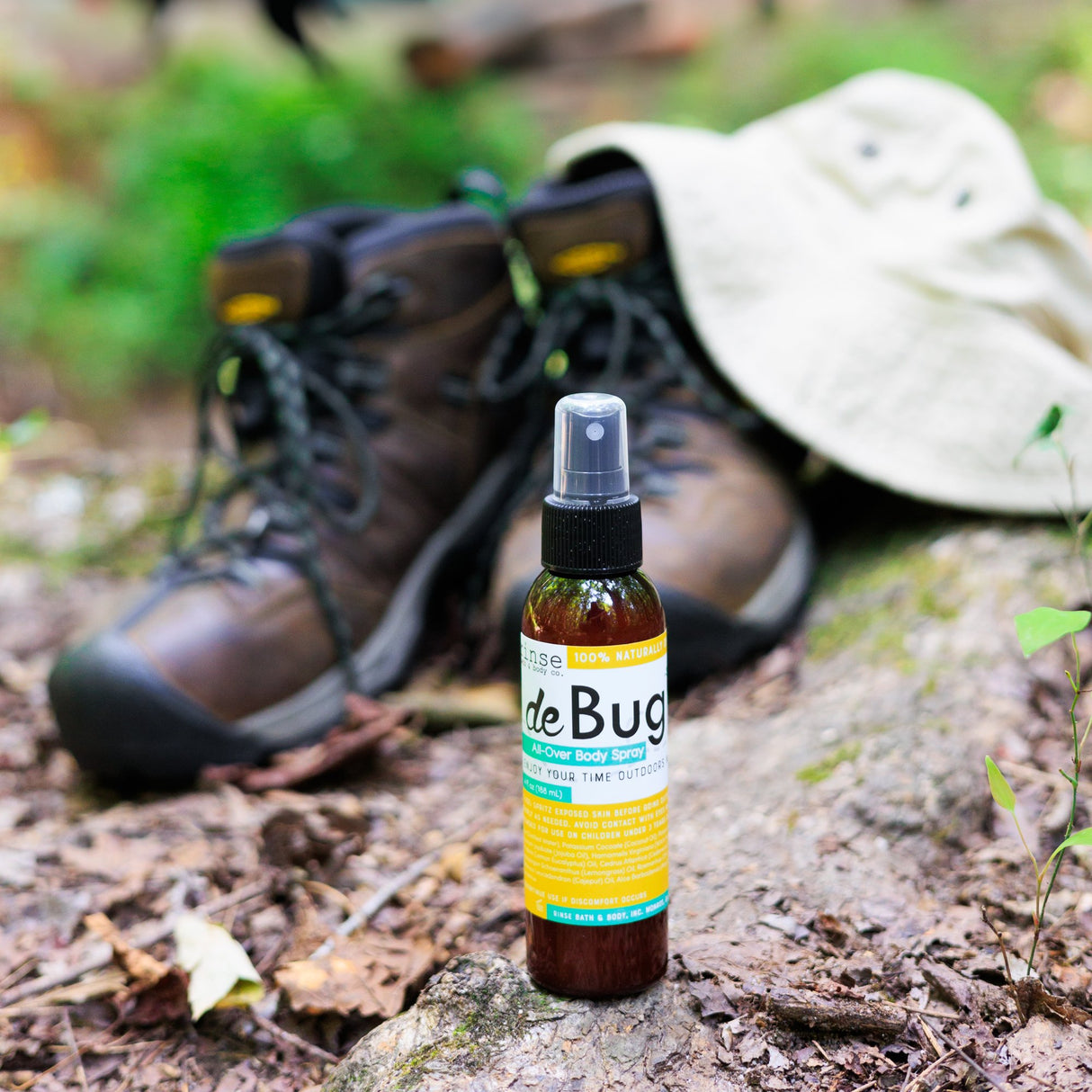 DeBug natural bug repellent spray beside hiking boots and a sun hat on a wooded trail