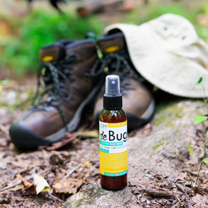 DeBug natural bug repellent spray beside hiking boots and a sun hat on a wooded trail
